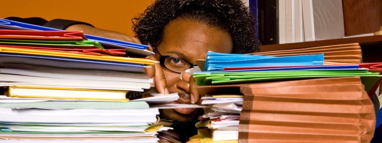 male buried in work looks between the two stacks of folders piled on top of his desk.