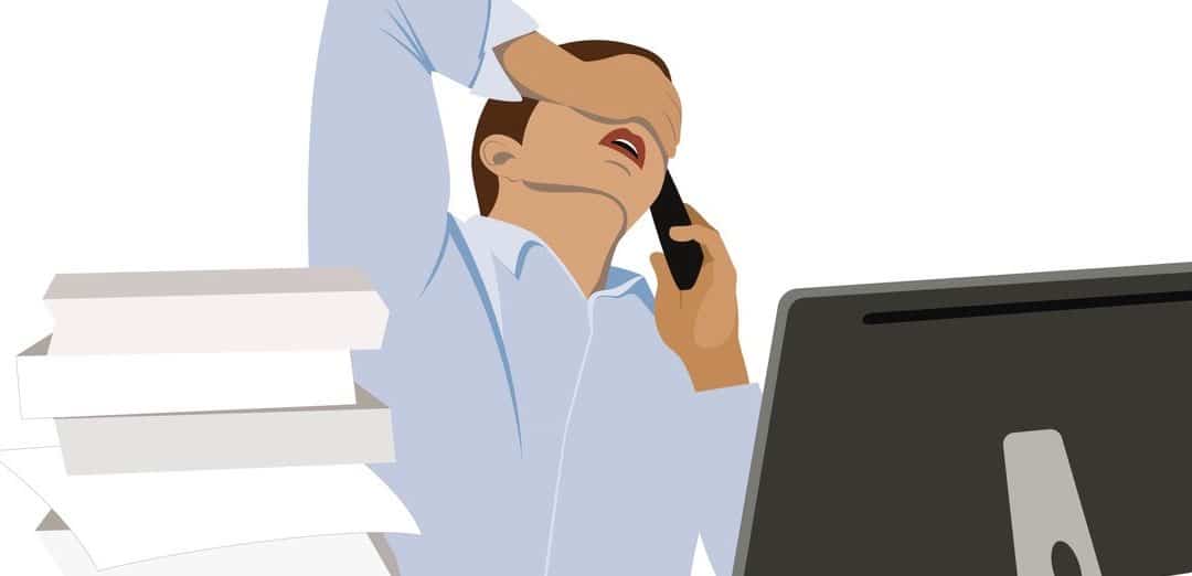 A young man sits at his desk holding his head and talking on his mobile phone