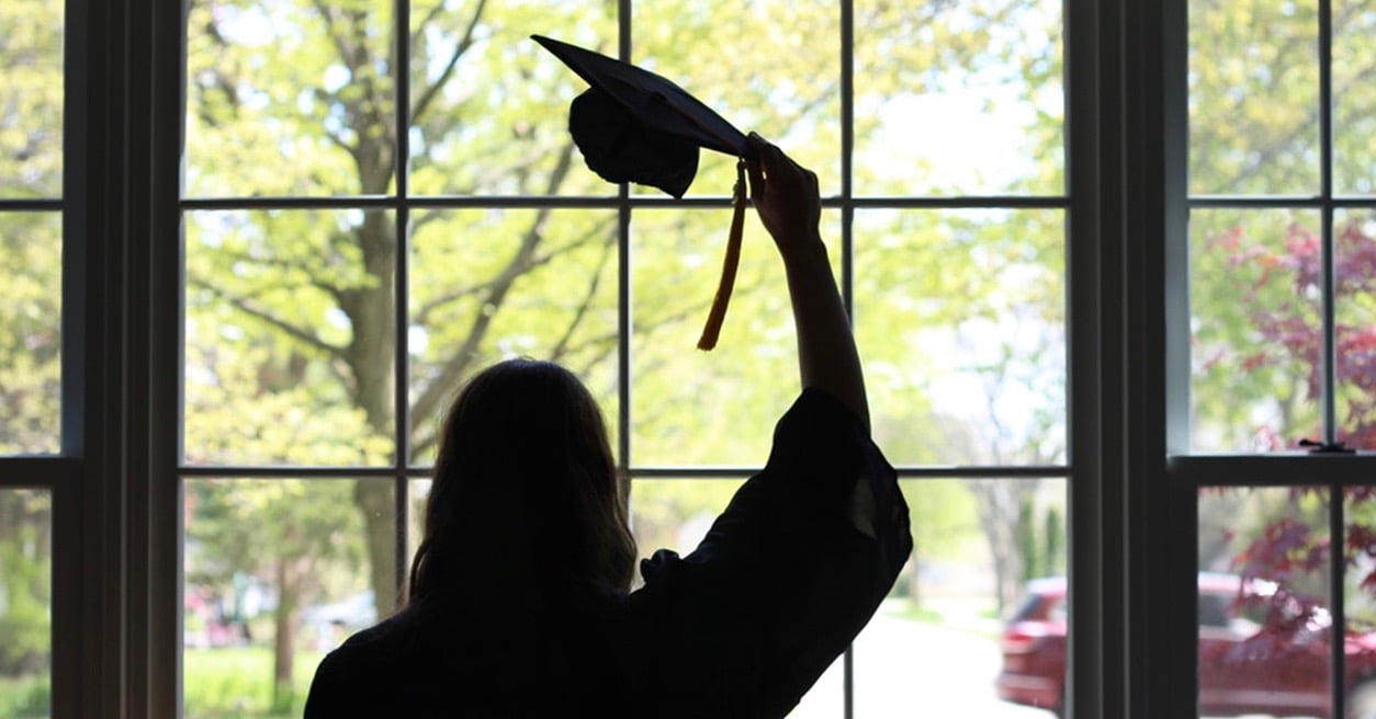 High school graduate removes her cap while staring out at the neighborhood, watching a car pass by during a virtual graduation ceremony.