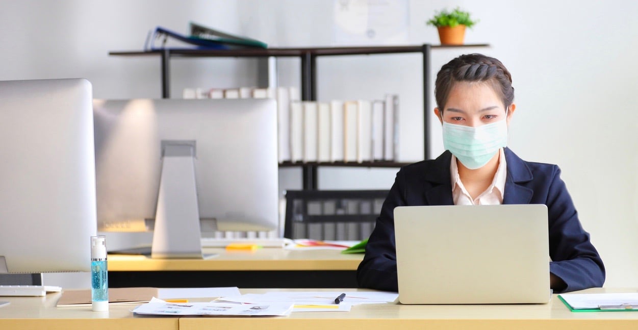 Female employee wearing medical face mask while working alone because of social distancing policy in the business office