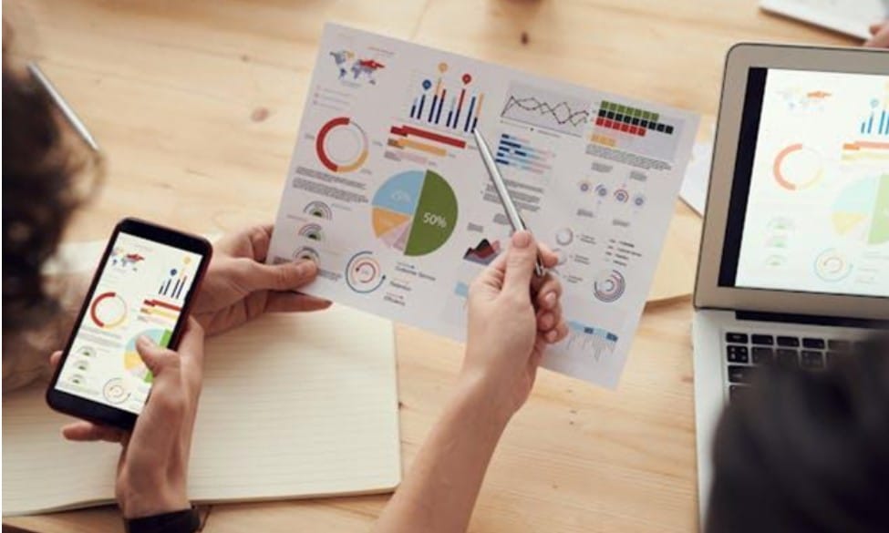 Two people looking at various charts on a piece of paper, phone, and laptop. 