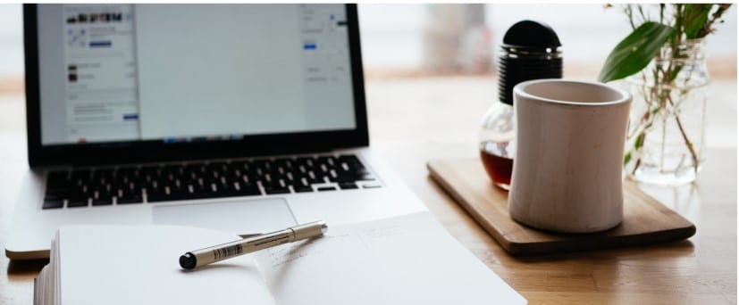 Open notebook with pen beside laptop and coffee cup on wooden desk.